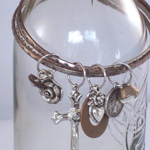 May include: A clear glass bottle with a silver-toned bangle bracelet around the neck. Dangling from the bracelet are several charms, including a cross, a coin, a bell, and a heart-shaped pendant. The charms are attached with small silver rings.