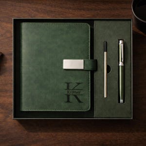 Peut inclure: Un ensemble de journal, stylo et crayon vert foncé avec une montre sur une surface en bois. Le journal a un fermoir argenté et le nom "Kilmer" gravé dessus. La montre a un bracelet en cuir marron.