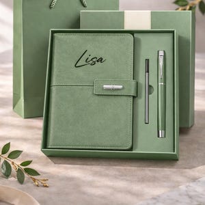 Peut inclure: Un coffret cadeau vert sauge comprenant un carnet avec "Lisa" en relief, un stylo et une recharge de stylo. L'ensemble est présenté dans une boîte assortie, avec un sac cadeau en arrière-plan. Le thème général est un cadeau personnalisé.