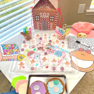 May include: A festive gingerbread house-shaped box with a red handle sits on a white table, surrounded by gingerbread-themed craft supplies. Includes felt cookies, gingerbread men, and candy canes. A fun, colorful craft kit.
