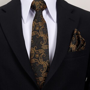 Op de afbeelding: Een zwart pak met een wit overhemd met kraag, een zwarte stropdas en een bijpassende pochet. De stropdas en pochet hebben een gouden paisley patroon. Het pak is een klassiek formeel ensemble.
