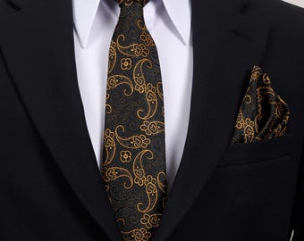 May include: A black suit with a white collared shirt, a black tie, and a matching pocket square. The tie and pocket square feature a gold paisley pattern. The suit is a classic formal wear ensemble.