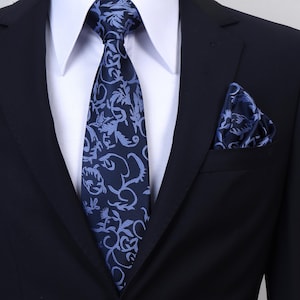 Könnte beinhalten: Eine dunkelmarineblaue Krawatte und ein passendes Einstecktuch mit einem hellblauen Blumen- und Rankenmuster. Das Set wird mit einem weißen Hemd mit Kragen und einem einfarbigen dunkelmarineblauen Sakko getragen, was ein formelles Erscheinungsbild ergibt.