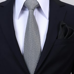 May include: A dark navy blue suit jacket worn over a crisp white collared shirt. A silver and black necktie features a repeating geometric dot pattern. A matching black pocket square with a subtle textured pattern is tucked into the breast pocket.