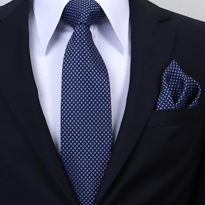 Può includere: Un blazer blu navy scuro indossato sopra una camicia bianca con colletto. Una cravatta e una pochette abbinata presentano un motivo ripetuto di piccoli rombi azzurri su uno sfondo blu navy, creando un aspetto strutturato e uniforme.