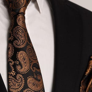 Op de afbeelding: Een zwarte stropdas met een paisley-patroon in goud. De stropdas heeft een klassiek ontwerp en is perfect om elke outfit een vleugje elegantie te geven.