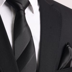 Könnte beinhalten: Nahaufnahme einer schwarzen Krawatte mit grauen Streifen, einem passenden Einstecktuch und einem schwarzen Anzug. Die Krawatte ist geknotet und über einem weißen Hemd mit Kragen getragen. Der Anzug ist maßgeschneidert.