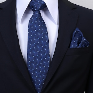 Peut inclure: Une cravate bleu foncé ornée d'un motif géométrique répétitif composé de petits carrés et de lignes diagonales bleu clair et blanc, accompagnée d'une pochette assortie, portée sur une chemise blanche à col et un blazer bleu marine uni.