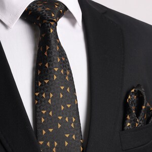 May include: A black tie with a geometric pattern of gold triangles. The tie is worn with a white shirt and a black suit jacket. A matching pocket square is also visible.