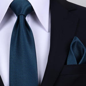 May include: A dark teal necktie and matching pocket square featuring a subtle, repeating chevron pattern. The necktie is tied in a knot and worn with a crisp white collared shirt and a dark navy blue blazer, creating a formal and coordinated ensemble.