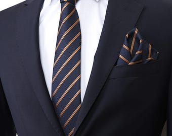 May include: A navy blue suit with a white dress shirt, a striped tie, and a matching pocket square. The tie and pocket square feature a pattern of gold stripes on a navy blue background. The suit jacket is buttoned.