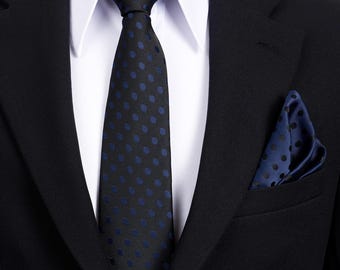 Puede incluir: Un traje negro con una camisa blanca, una corbata negra con lunares azules y un pañuelo de bolsillo a juego. La corbata está anudada y el pañuelo está cuidadosamente doblado en el bolsillo del pecho del traje.