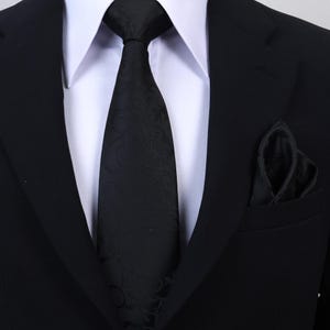 Könnte beinhalten: Eine schwarze Krawatte mit einem dezenten, tonalen Blumenmuster, kombiniert mit einem passenden schwarzen Einstecktuch. Das Set wird zu einem weißen Hemd mit Kragen und einem schwarzen Sakko getragen, was einen formellen, monochromen Look ergibt.