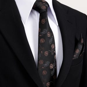 Peut inclure: Une cravate et une pochette noires avec un motif floral répété marron et gris. La cravate est nouée et portée avec une chemise blanche et une veste de costume noire.