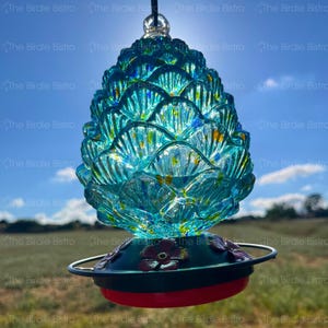 May include: A turquoise glass hummingbird feeder shaped like a pinecone. The feeder has a red base, a dark green top with floral accents, and a metal perch. The background shows a blue sky and green field.