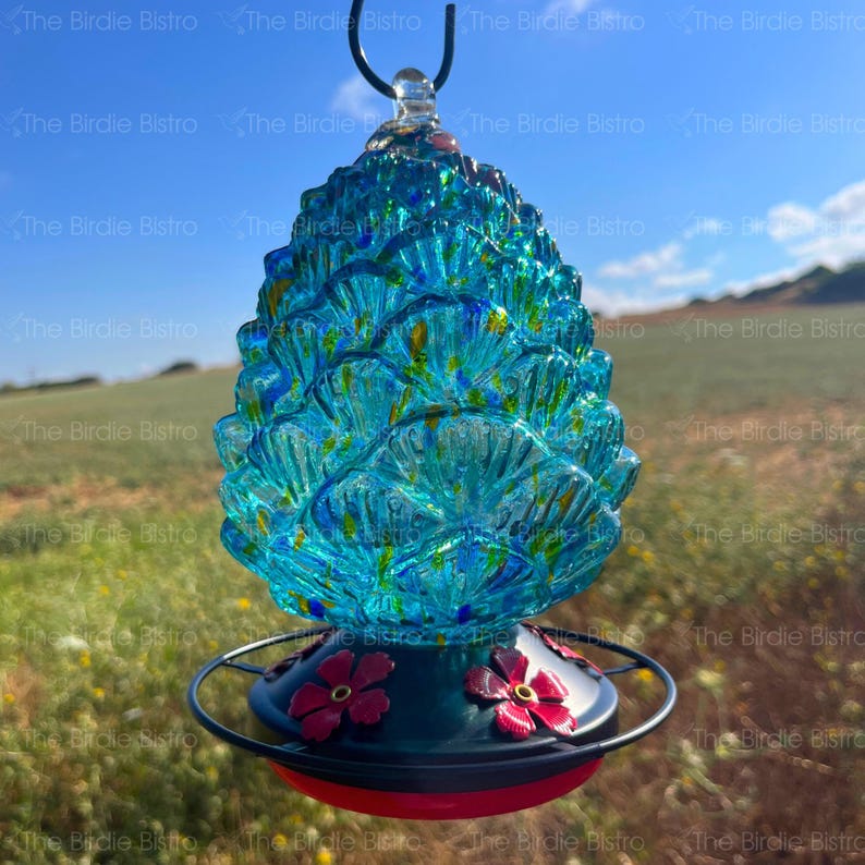 May include: A turquoise glass hummingbird feeder with a pinecone design. The feeder has a black metal base with red flower accents and a red feeding port. The text "The Birdie Bistro" is repeated across the image.
