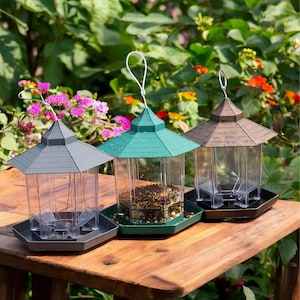 May include: Three bird feeders with clear plastic cages and different colored roofs. The feeders are on a wooden table. The feeder on the left has a gray roof, the middle feeder has a green roof, and the right feeder has a brown roof.