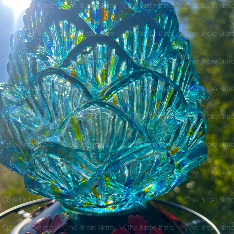 May include: A close-up of a turquoise glass bird feeder with a textured, layered design. The feeder has a flower-like shape with yellow accents. The words "The Birdie Bistro" are visible in the background.