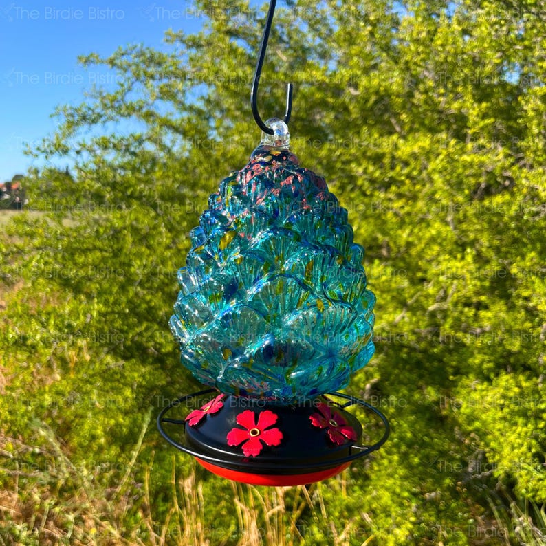 May include: A decorative hummingbird feeder with a blue, textured glass body shaped like a pinecone. The feeder has a black base with red flower accents and a red rim. It hangs from a black hook.
