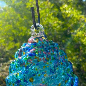 May include: A colorful, hand-blown glass bird feeder with a textured surface. The feeder features shades of blue, green, red, and yellow, with a clear glass loop for hanging. A black cord is attached to the loop. The Birdie Bistro is printed on the image.