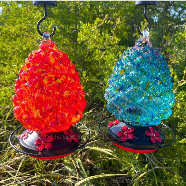 May include: Two decorative glass hummingbird feeders. One is red and orange, the other is blue and turquoise. Both have a black base with red flower accents and a metal hanger. The feeders are designed to attract hummingbirds.