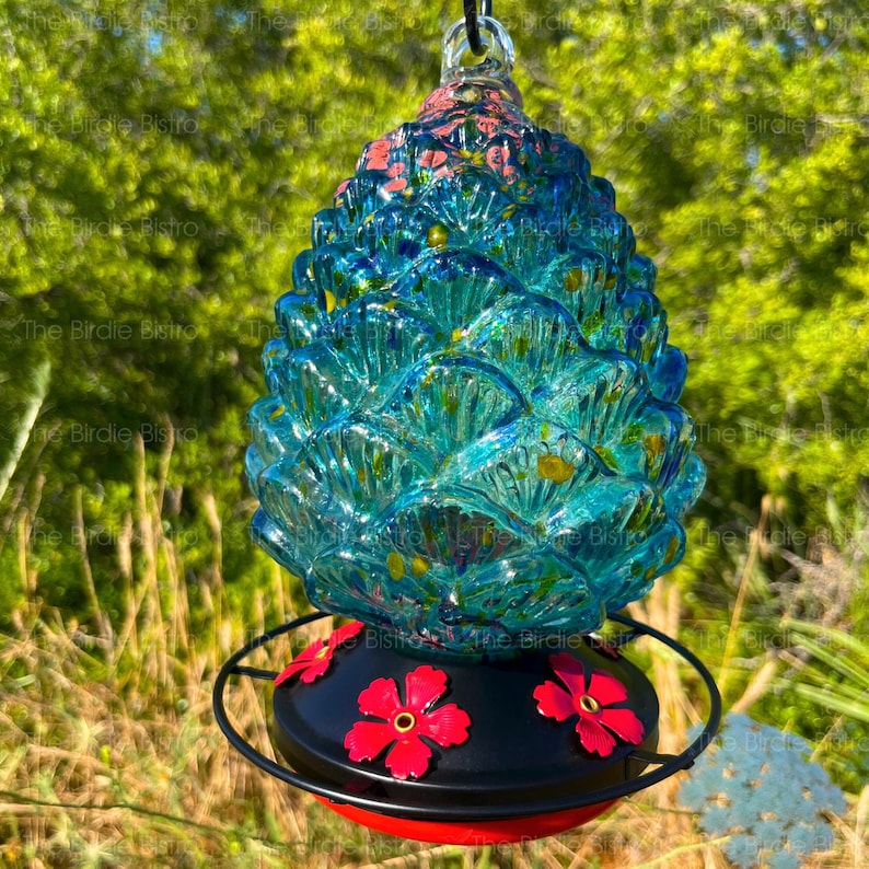 May include: A blue glass hummingbird feeder shaped like a pinecone, with red hibiscus flower accents. The feeder has a black base with a red rim and a metal hanging loop. The background is blurred green foliage.