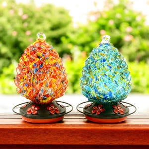 Puede incluir: Dos comederos de colibrí de vidrio con forma de piñas. Un comedero es azul con motas blancas y amarillas, el otro es naranja con motas rojas y amarillas. Ambos comederos tienen bases de metal negro con detalles de flores rojas.