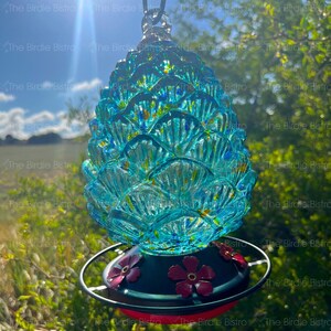 May include: A turquoise glass hummingbird feeder shaped like a pineapple. The feeder has a black metal base with red accents and decorative red flowers. The feeder is hanging outside with a bright blue sky in the background.