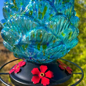 May include: A decorative hummingbird feeder with a vibrant, textured glass reservoir in shades of turquoise and blue. The base is black with bright red flower accents. A metal ring surrounds the base.