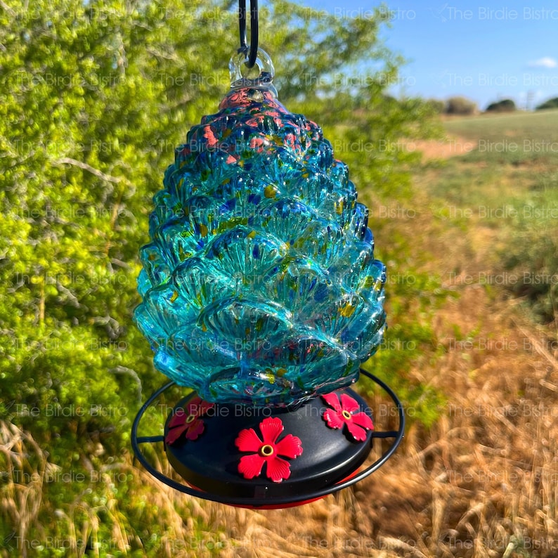 May include: A hanging hummingbird feeder with a blue glass body and a black base. The feeder has a textured, cone-like shape with red flower accents on the base. The feeder is suspended by a black cord.