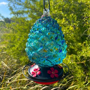 May include: A decorative glass bird feeder in the shape of a pinecone. The feeder is turquoise and blue with yellow accents. The base is black with red flowers and a red rim. A black wire hangs from the top.