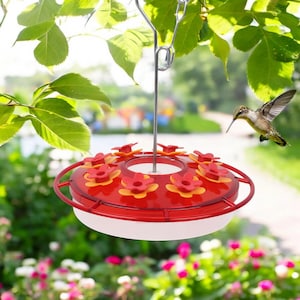 Puede incluir: Comedero para colibríes rojo con una base blanca y seis perchas en forma de flor roja. El comedero está colgado de un gancho de metal y un colibrí vuela hacia él.