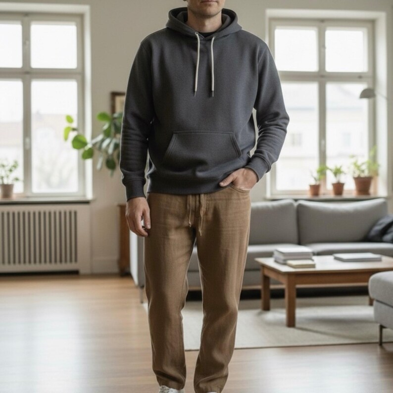 Peut inclure: Une personne porte un sweat &agrave; capuche gris fonc&eacute; avec une poche avant et des cordons blancs, associ&eacute; &agrave; un pantalon beige. La personne se tient dans un salon bien &eacute;clair&eacute; avec un canap&eacute; et des fen&ecirc;tres.
