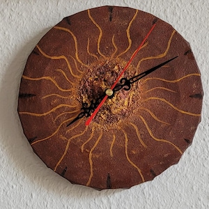 May include: A round, brown wall clock with a sunburst design. The clock face features a textured center with gold and red accents, and gold lines radiating outwards. The clock hands are black with a red second hand.