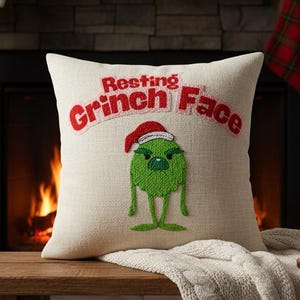 May include: A cream-colored square pillow with the text "Resting Grinch Face" in red embroidery. The pillow features an embroidered green Grinch character wearing a Santa hat. The pillow is on a wooden bench in front of a fireplace.
