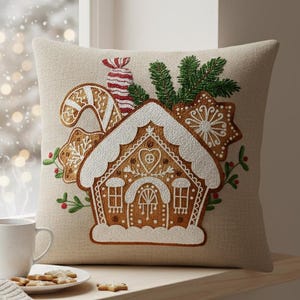 May include: Beige square cushion with an embroidered gingerbread house design. The cushion features a gingerbread house, candy cane, and star-shaped biscuits. Green sprigs and red accents add colour to the festive design. A white mug and biscuits sit on a wooden windowsill.