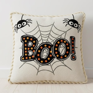 May include: Square Halloween-themed pillow with a cream-colored background and a rope trim. The pillow features a spider web design with two black spiders and the word "BOO!" in black letters with orange polka dots.