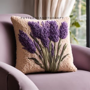 May include: A square, beige throw pillow with a textured surface. The pillow features a detailed, embroidered design of purple lavender flowers with green stems and leaves. The text "PATTERNSANDTHERAPY" is printed on the top of the pillow.