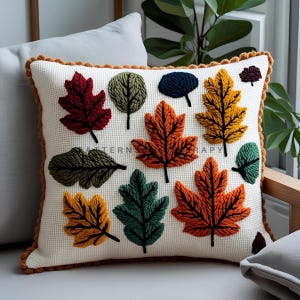 May include: Square decorative pillow with a cream-colored background and a brown border. The pillow features embroidered leaves in various colors, including red, orange, yellow, green, and navy blue. The text "PATTERN THERAPY" is visible on the pillow.