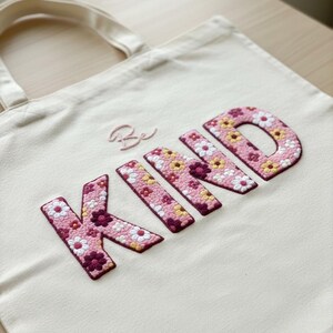 Op de afbeelding: Crèmekleurige draagtas met de geborduurde woorden "Be KIND". De letters zijn gevuld met een bloemenpatroon in roze, wit en geel. De tas heeft een eenvoudig ontwerp.