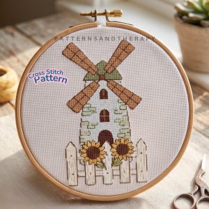 May include: A cross-stitch pattern of a windmill with brown blades, a white stone building, and a picket fence with sunflowers. The text "Cross Stitch Pattern" is on the left side, and "PATTERNSANDTHERAPY" is at the top. The embroidery is in a wooden hoop.