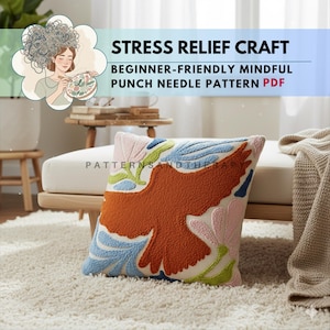 Op de afbeelding: Een decoratief kussen met een punch needle-ontwerp met een oranje vogel met uitgespreide vleugels. Het kussen heeft een witte achtergrond met blauwe, roze en groene bloemaccenten. De tekst op de afbeelding luidt "STRESS RELIEF CRAFT" en "BEGINNER-FRIENDLY MINDFUL PUNCH NEEDLE PATTERN PDF."