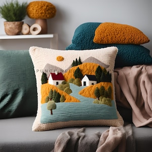 May include: A white throw pillow with a punch needle embroidery design of a small village with houses, trees, and a river. The pillow is on a gray couch with other pillows in teal and mustard yellow.