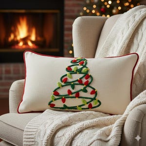 May include: Cream-colored rectangular pillow with a Christmas tree design. The tree is made of green yarn with red and green bulb accents. The pillow has a red border and is placed on a beige chair with a white blanket.