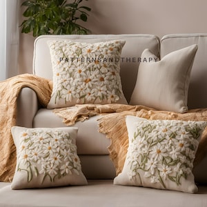 May include: Four throw pillows with white embroidered flowers on a beige couch. The pillows are arranged in a decorative way, with two floral pillows in the front and two plain beige pillows in the back. The couch is covered in a beige fabric and has a brown throw blanket draped over it.