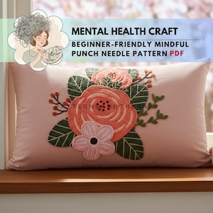 Könnte beinhalten: Ein rechteckiges, hellrosa Kissen mit floralem Design. Das Design zeigt eine große, korallenfarbene Rose, eine kleinere rosa Blume und grüne Blätter. Der Text "MENTAL HEALTH CRAFT BEGINNER-FRIENDLY MINDFUL PUNCH NEEDLE PATTERN PDF" wird angezeigt.