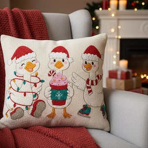 Op de afbeelding: Een decoratief kussen met drie ganzen met kerstmutsen en kerstaccessoires. Eén gans is omwikkeld met lichtjes, een andere houdt een drankje vast en de derde draagt schaatsen. Het kussen ligt op een stoel met een rode deken.