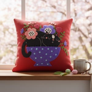 Könnte beinhalten: Ein rotes Zierkissen mit einem gestickten Design. Das Design zeigt eine schwarze Katze, die in einer lila Schale mit Punkten schläft, umgeben von bunten Blumen. Das Kissen liegt auf einer Holzoberfläche in der Nähe eines Fensters.