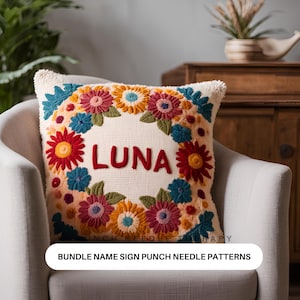 May include: A white pillow with a floral embroidery design in shades of red, orange, yellow, blue, and green. The name "LUNA" is embroidered in red in the center of the pillow. The pillow is on a white couch.