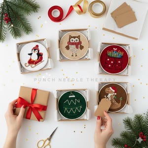 Könnte beinhalten: Eine Sammlung von Weihnachts-Stickrahmen in kleinen Pappschachteln. Designs umfassen einen Pinguin auf einem Schlitten, eine Eule mit Schal, eine Geschenktüte, eine grüne Christbaumkugel und einen Dinosaurier mit Weihnachtsmütze. Der Text "PUNCH NEEDLE THERAPY" ist sichtbar.
